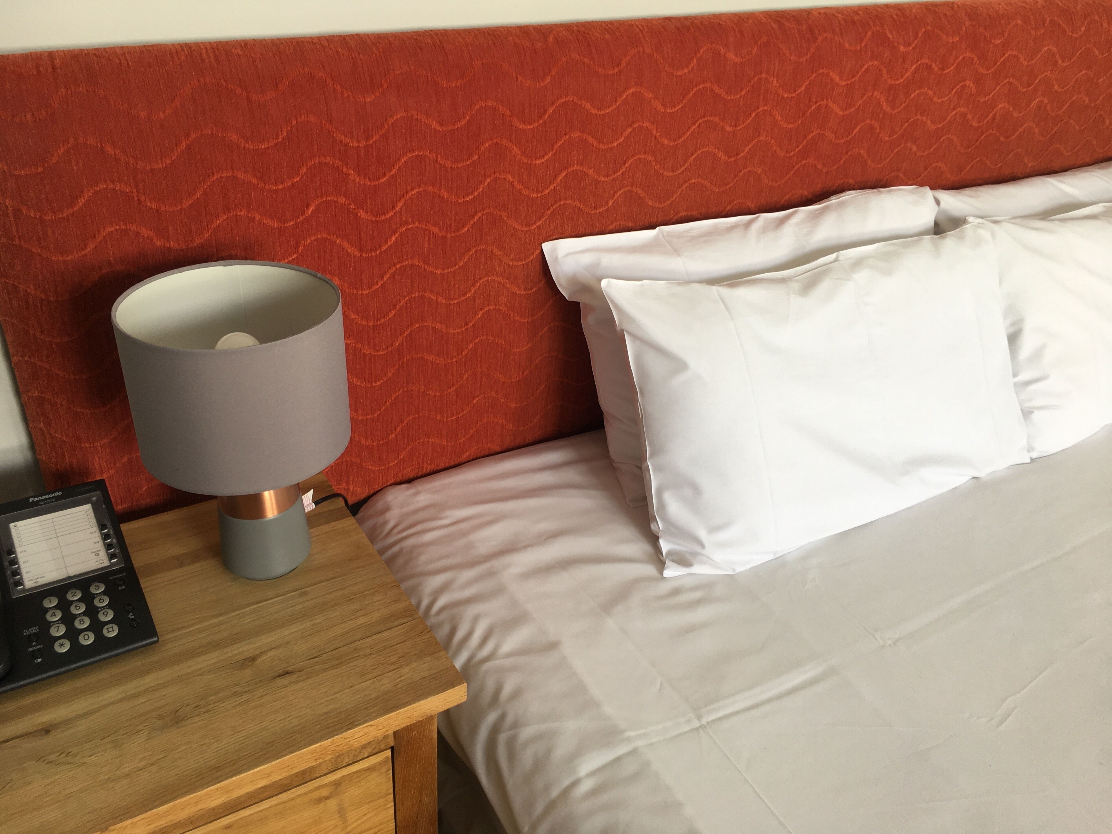A neatly made bed with white linens, a bedside lamp, and a small bedside table with a telephone.