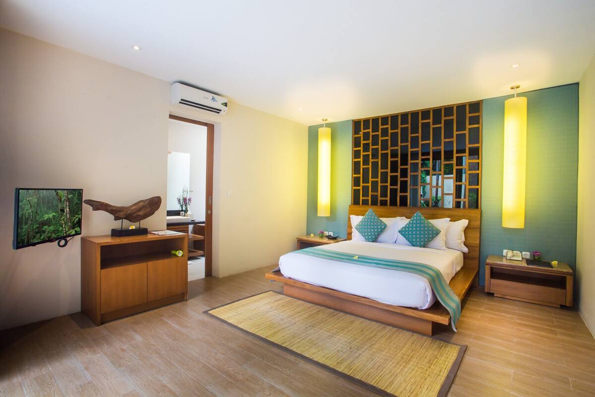 Quarto de hotel moderno com cama de madeira, mesa de cabeceira, escrivaninha e televisão presa à parede.