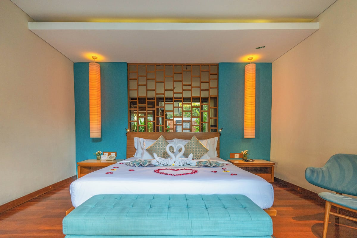Quarto de hotel com uma cama grande, cabeceira de madeira, duas mesinhas de cabeceira, uma cadeira e um painel decorativo na parede.