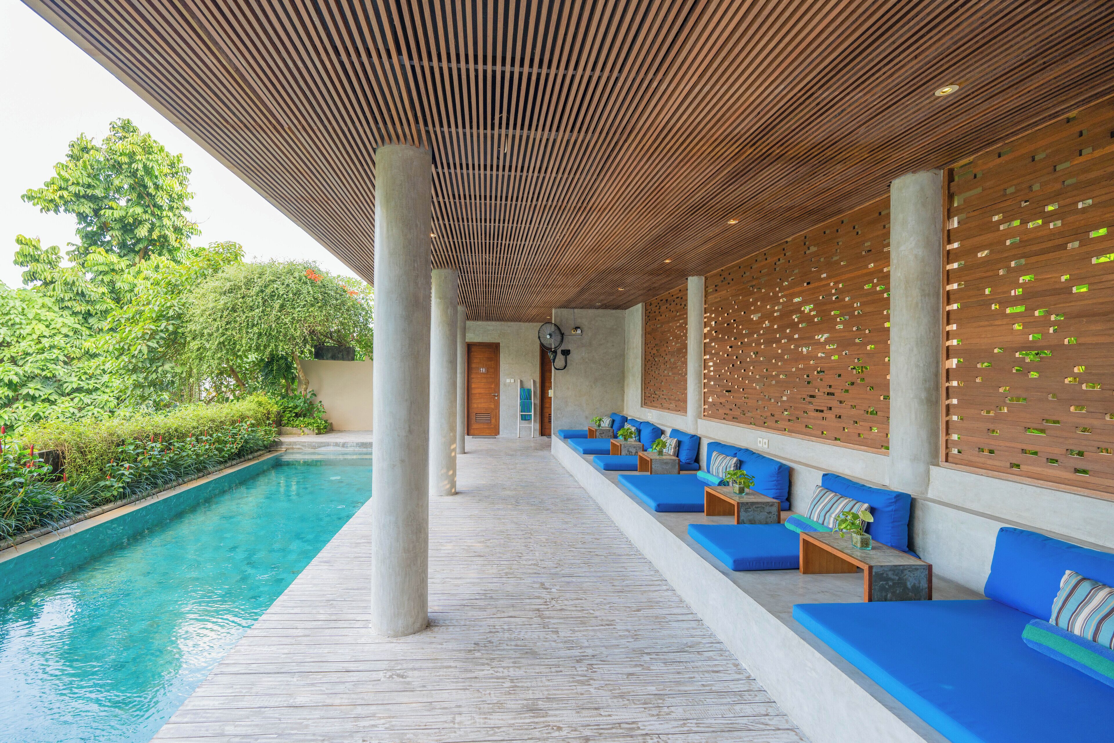 A poolside area with wooden slatted ceiling, blue cushioned seating, and a concrete floor.