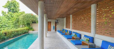 A poolside area with wooden slatted ceiling, blue cushioned seating, and a concrete floor.