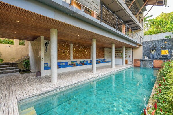 A swimming pool with a wooden deck, seating areas, and a stone wall with plants. - Maca Villas & Residence, Umalas (Kerobokan)