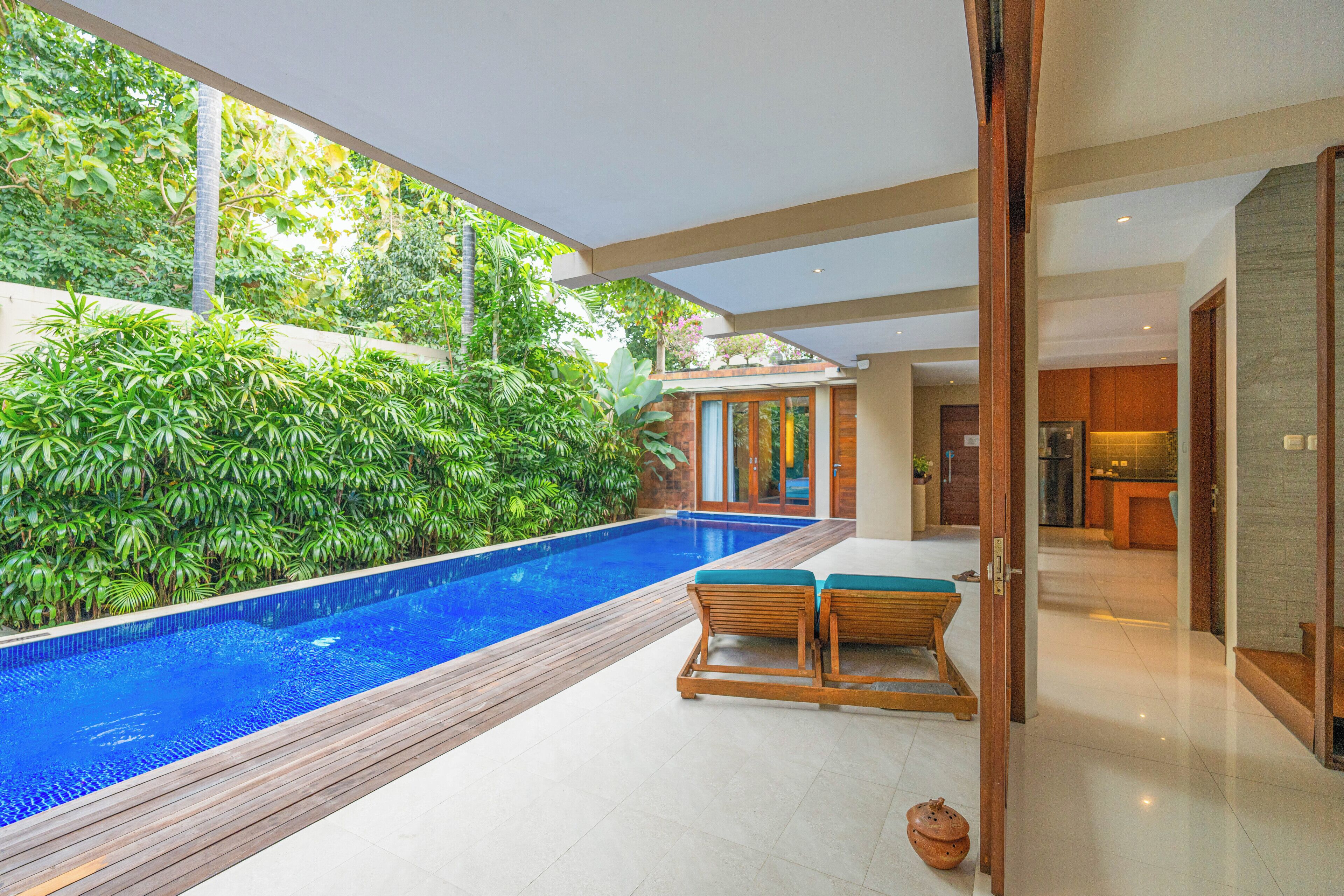 Un área de piscina interior moderna con una terraza de madera, dos sillones de madera y una exuberante pared verde.