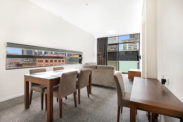 A modern hotel room with a dining area, a sofa, and a large window offering a city view.