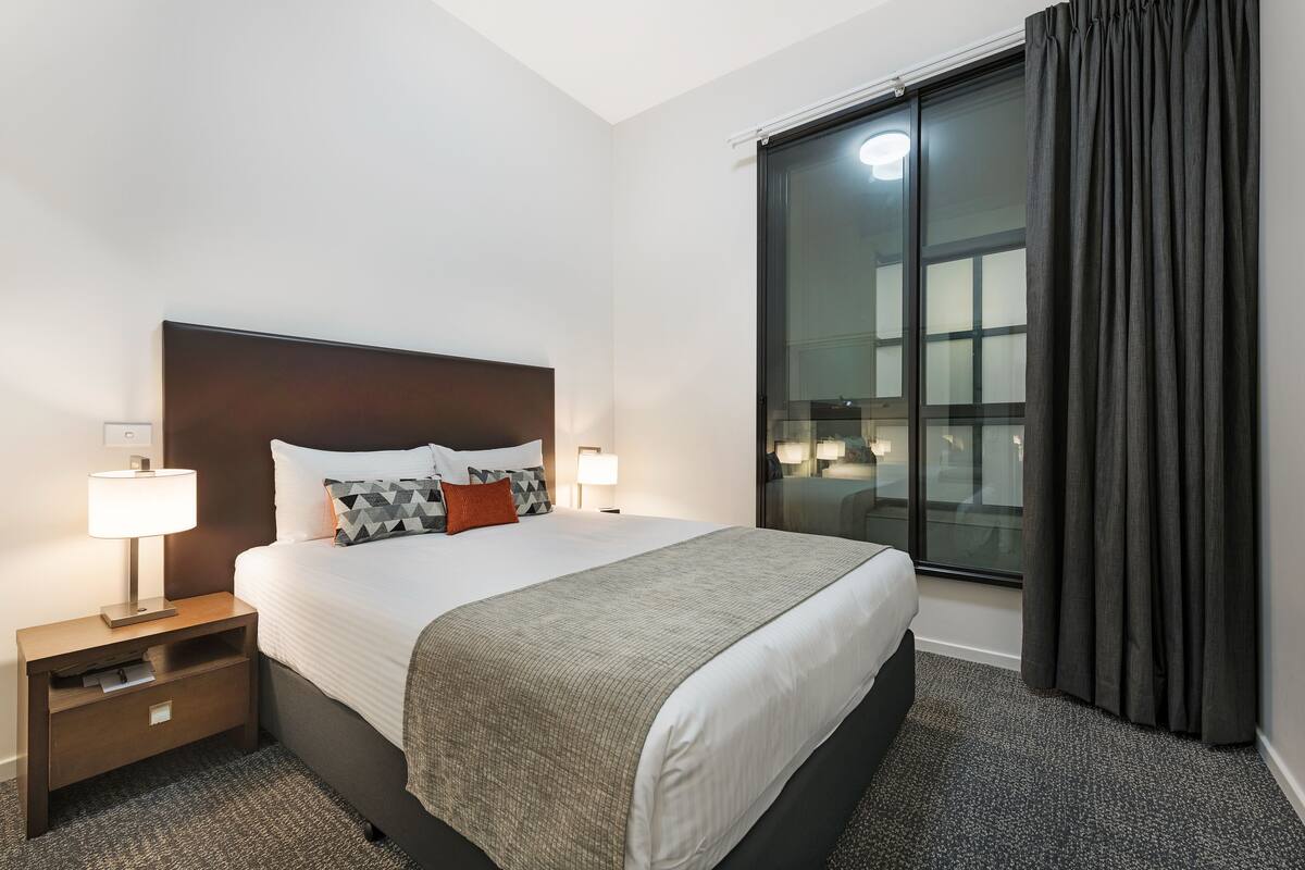 A hotel room with a large bed, two bedside lamps, a nightstand, a window with curtains, and a view of the city.