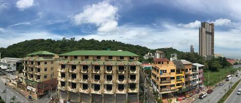A multi-story building with balconies, a green roof, and a view of a busy street and a hill with trees.