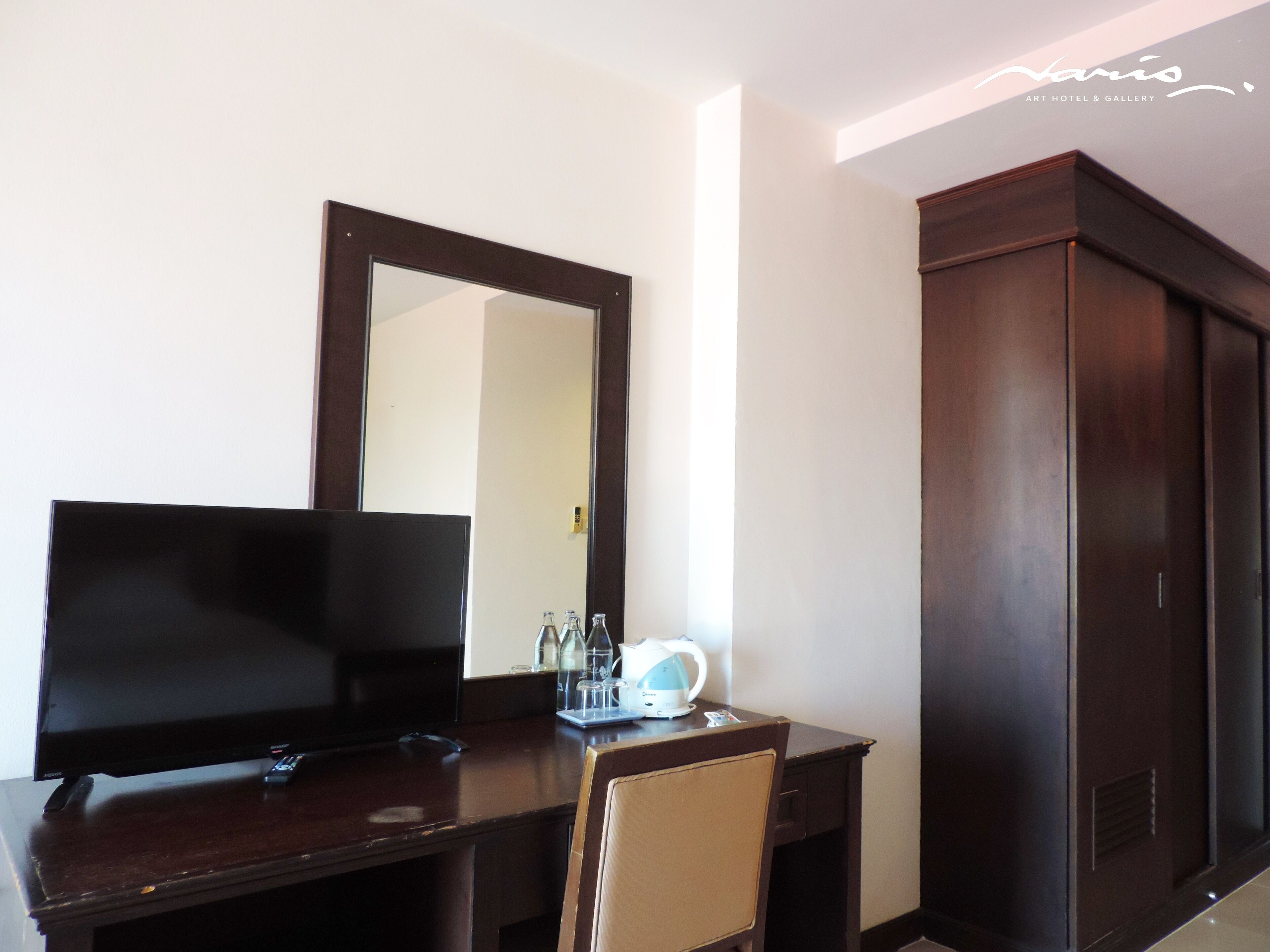 A hotel room with a dark wooden desk, a flat-screen TV, a mirror, a small table with a kettle, and a wardrobe.
