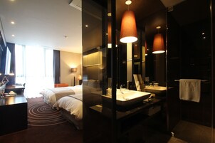 A modern hotel room with a large bed, a desk, a bathroom with a sink and mirror, and large windows.