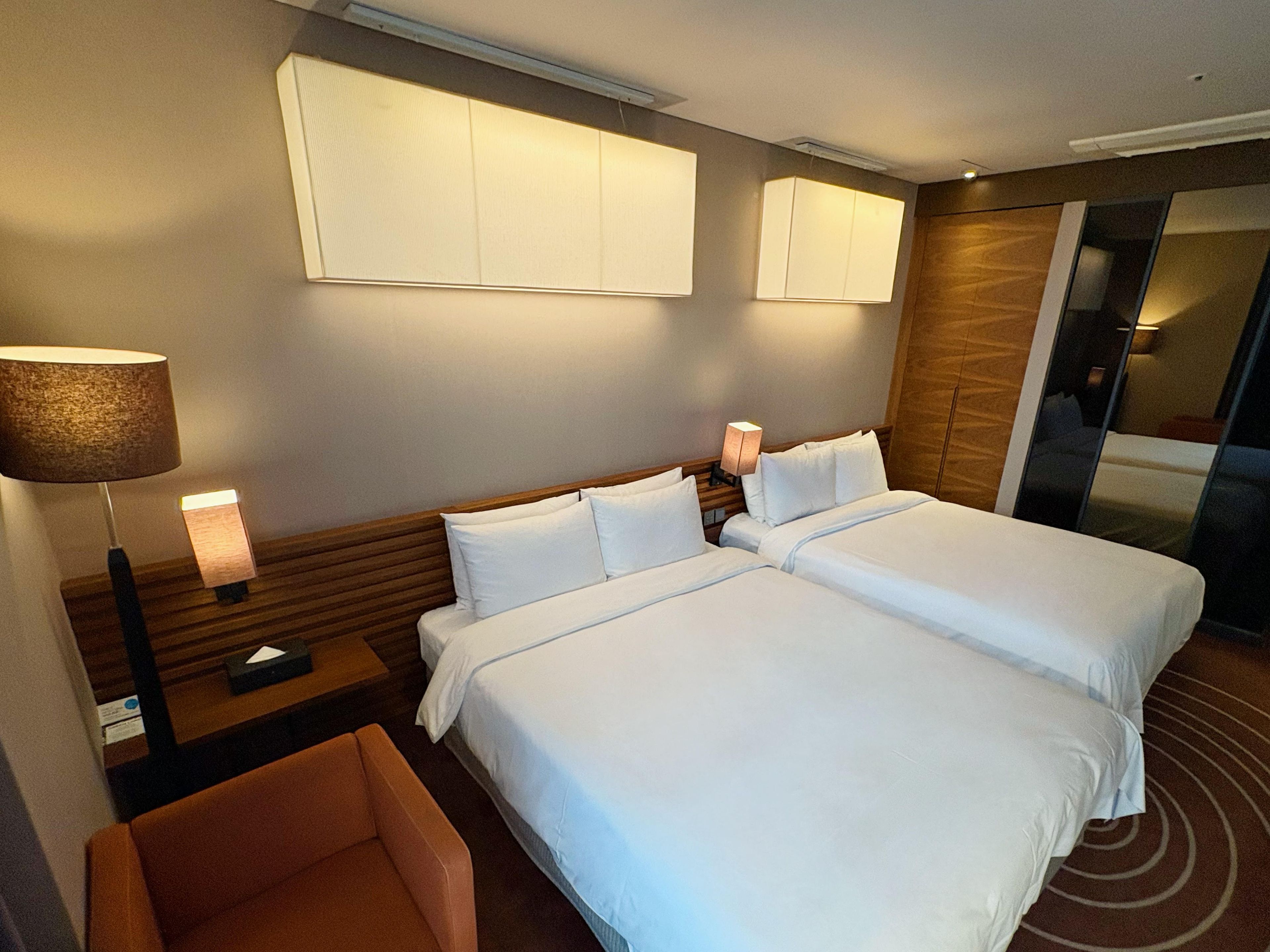 Quarto de hotel com duas camas, uma mesa de cabeceira, uma luminária e um espelho.