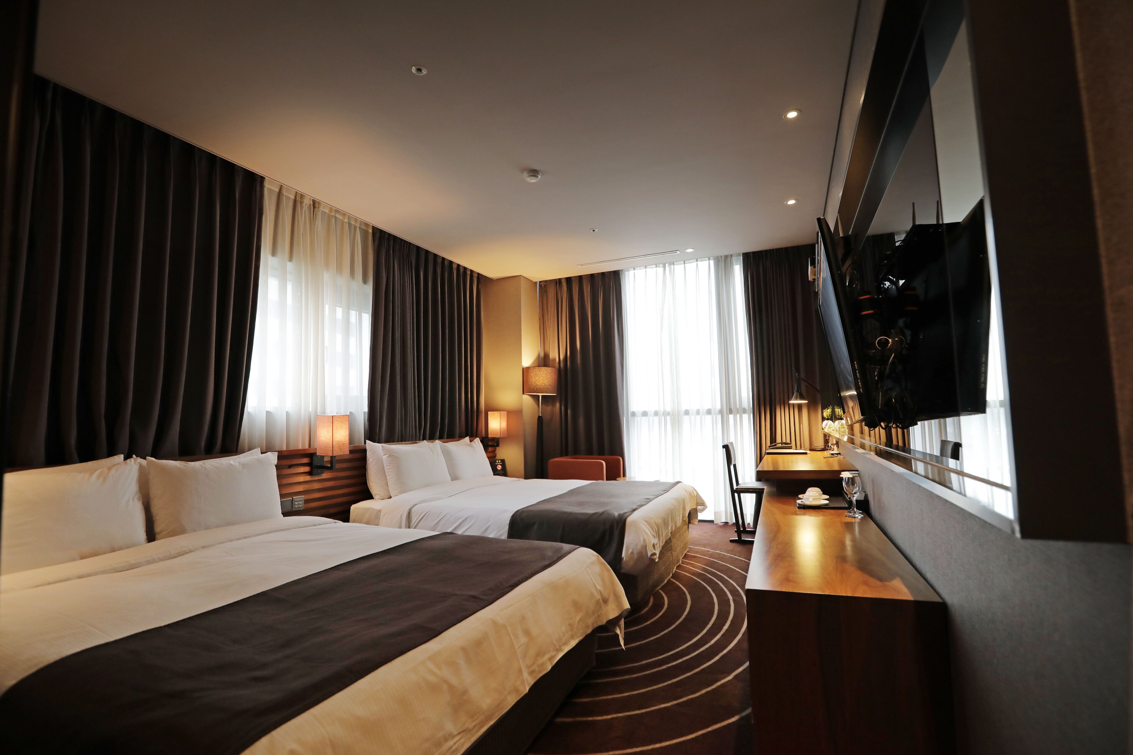 A hotel room with two beds, a desk, a TV, and large windows with curtains.