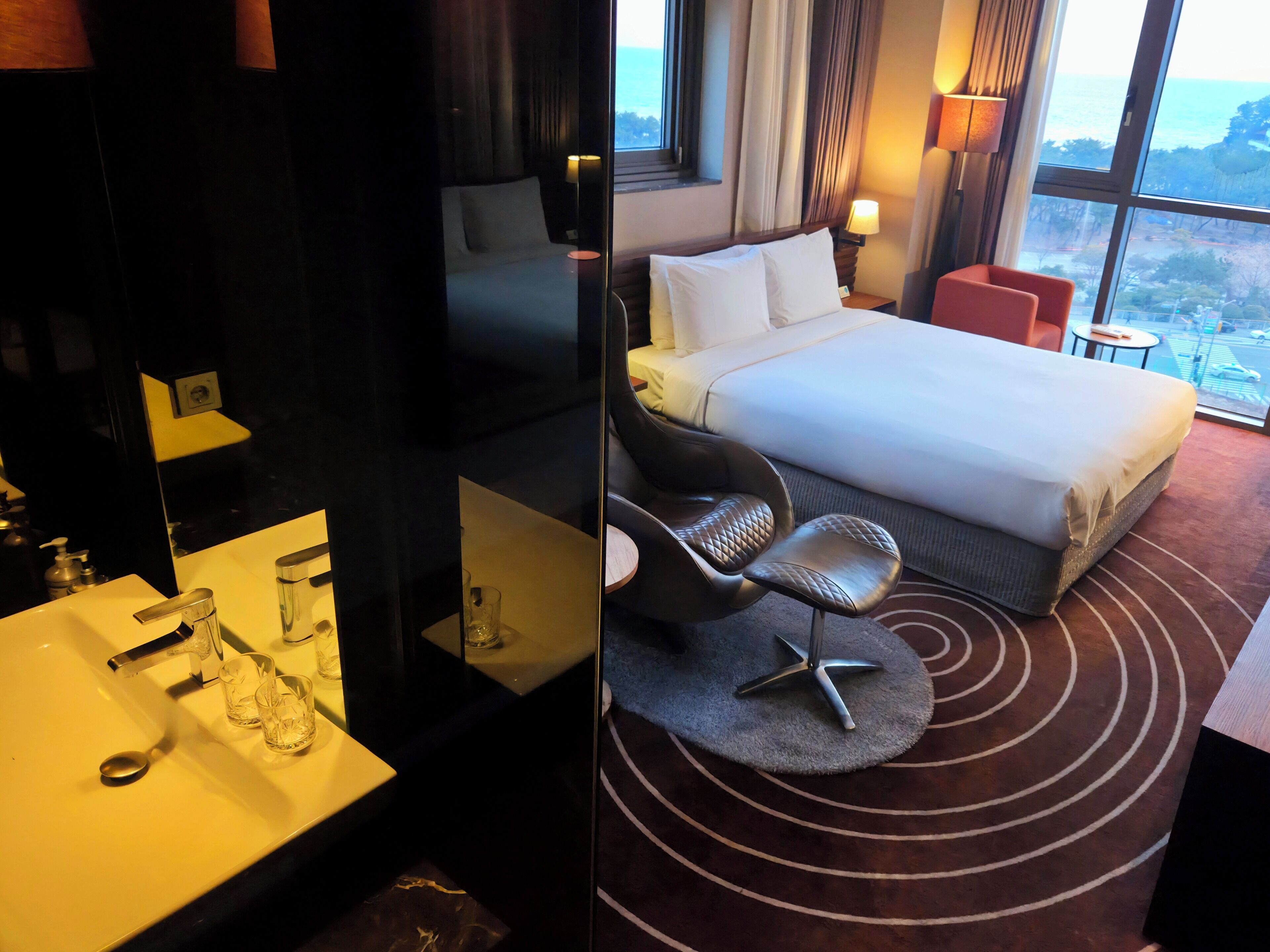 Habitación de hotel con cama, escritorio con espejo, lavamanos con vasos y vista al mar a través de amplias ventanas.