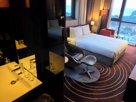 Habitación de hotel con cama, escritorio con espejo, lavamanos con vasos y vista al mar a través de amplias ventanas.