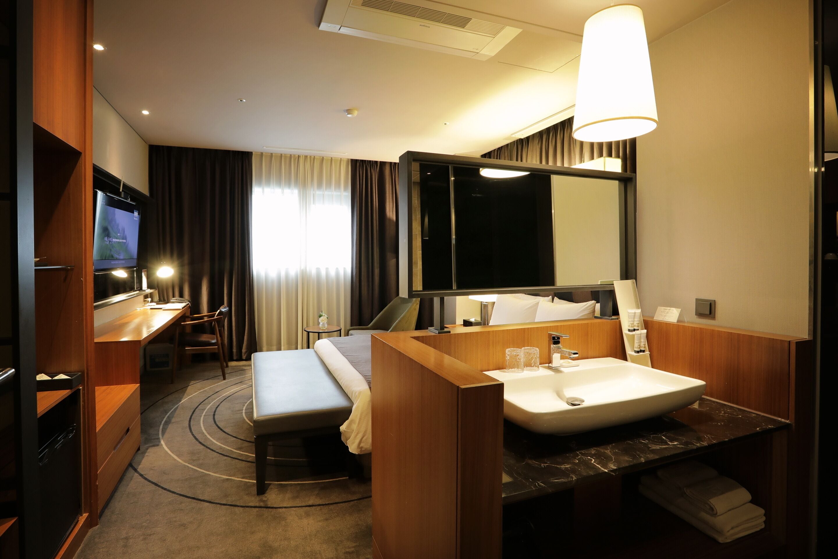 A hotel room with a bed, a desk, a TV, a large mirror, and a bathroom with a sink and towel rack.