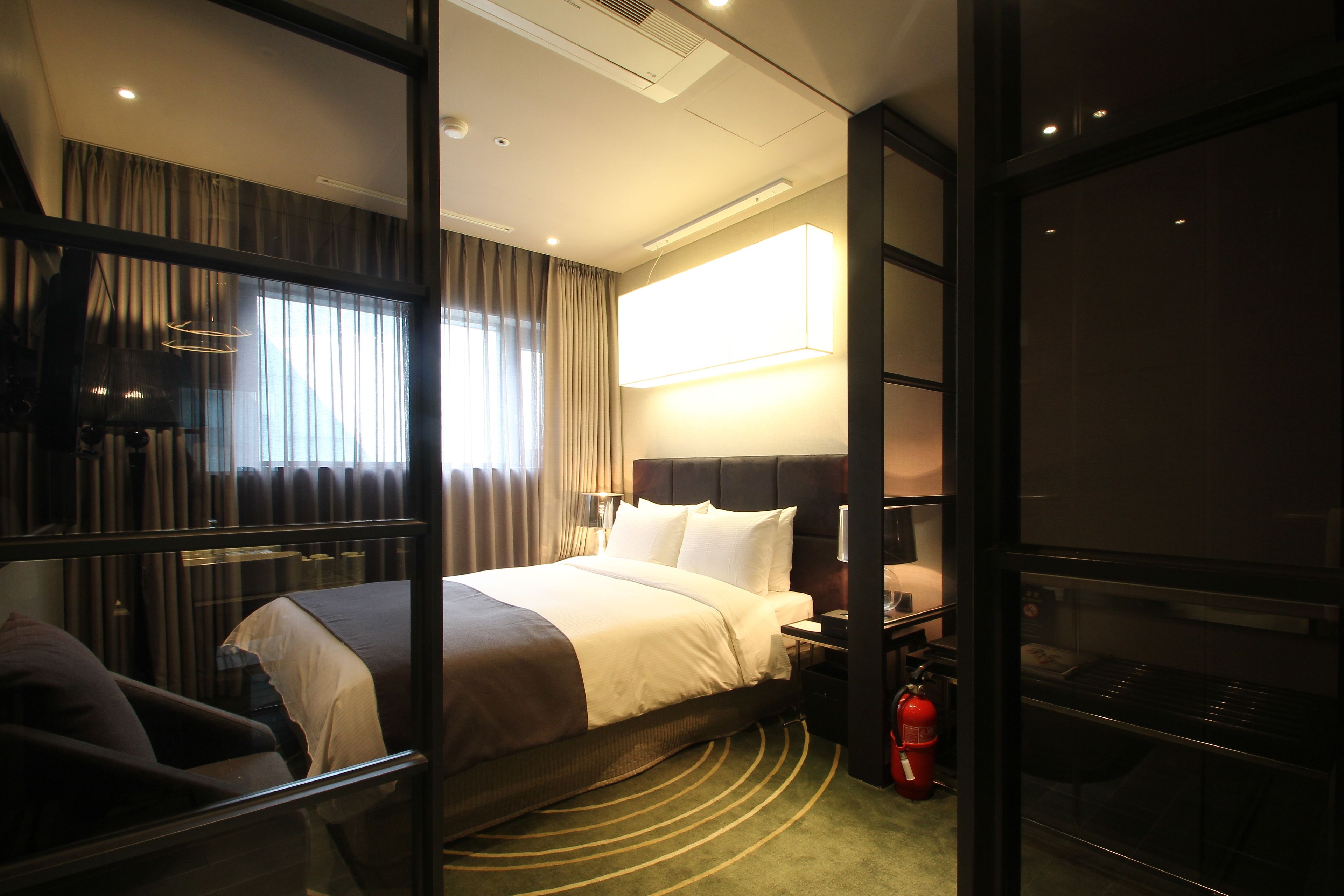 A hotel room with a large bed, a bedside table, a lamp, a fire extinguisher, and a view of the outside through the window.