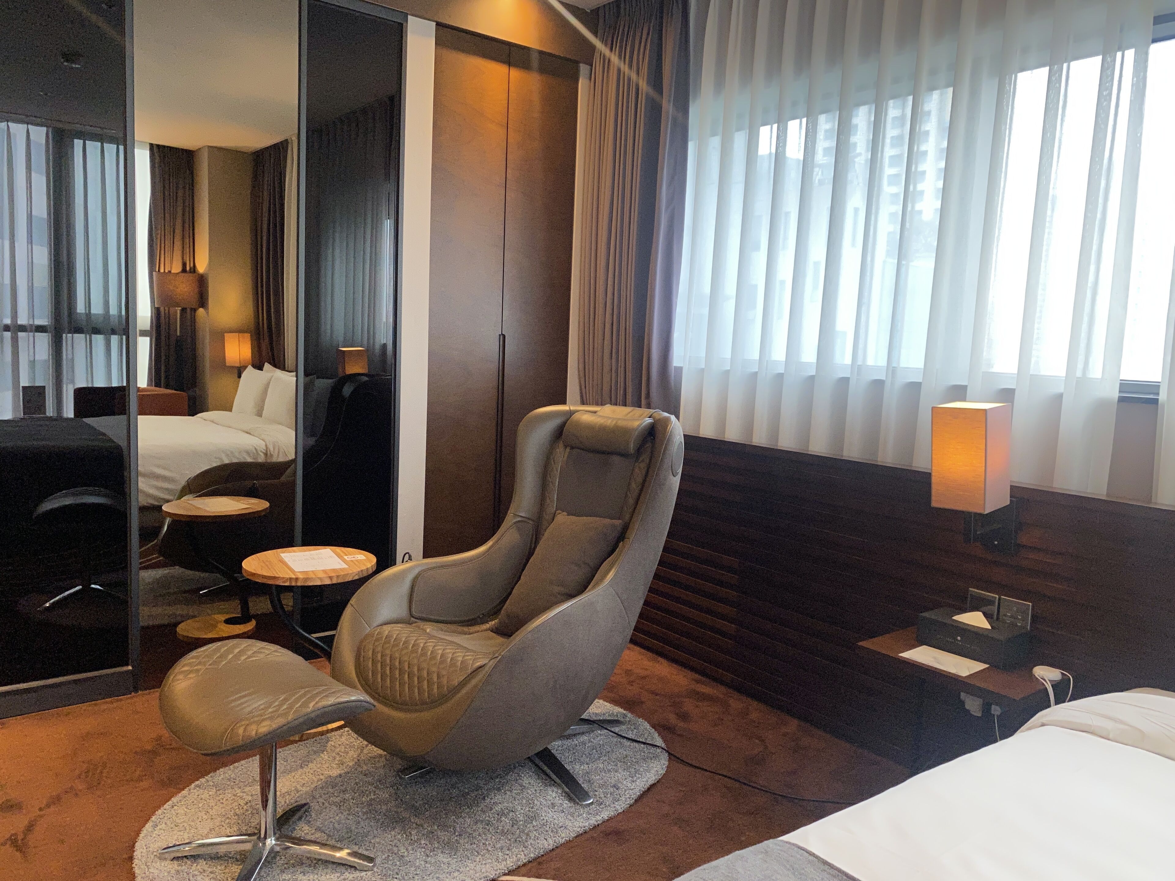 A modern hotel room with a large bed, a reclining chair, a small table, and a lamp.