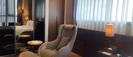 A modern hotel room with a large bed, a reclining chair, a small table, and a lamp.