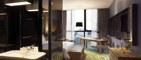 Una habitación de hotel moderna con una cama grande, un área de descanso con una mesa y sillas, un ventanal con cortinas, un televisor de pantalla plana y un baño con lavamanos y espejo.