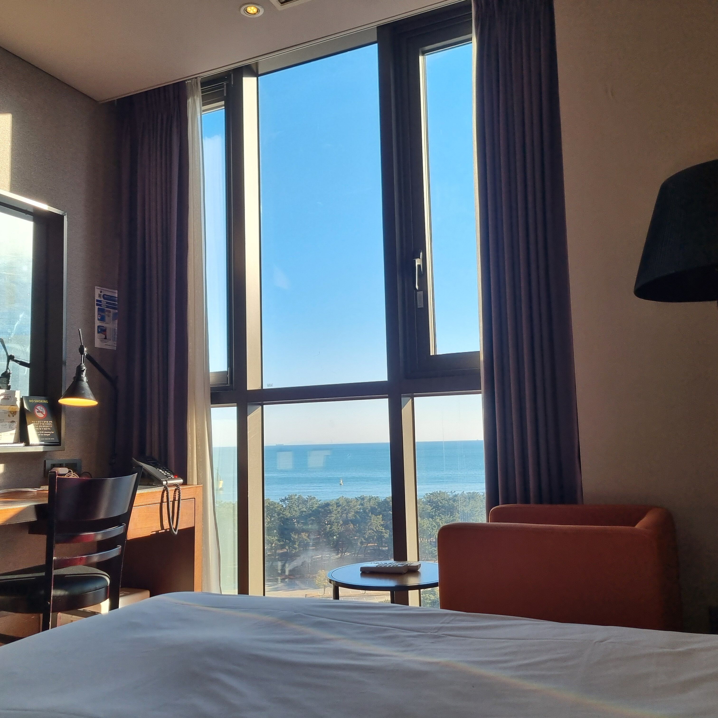 Et hotelværelse med et stort vindue, en seng, et skrivebord med lampe og en stol.