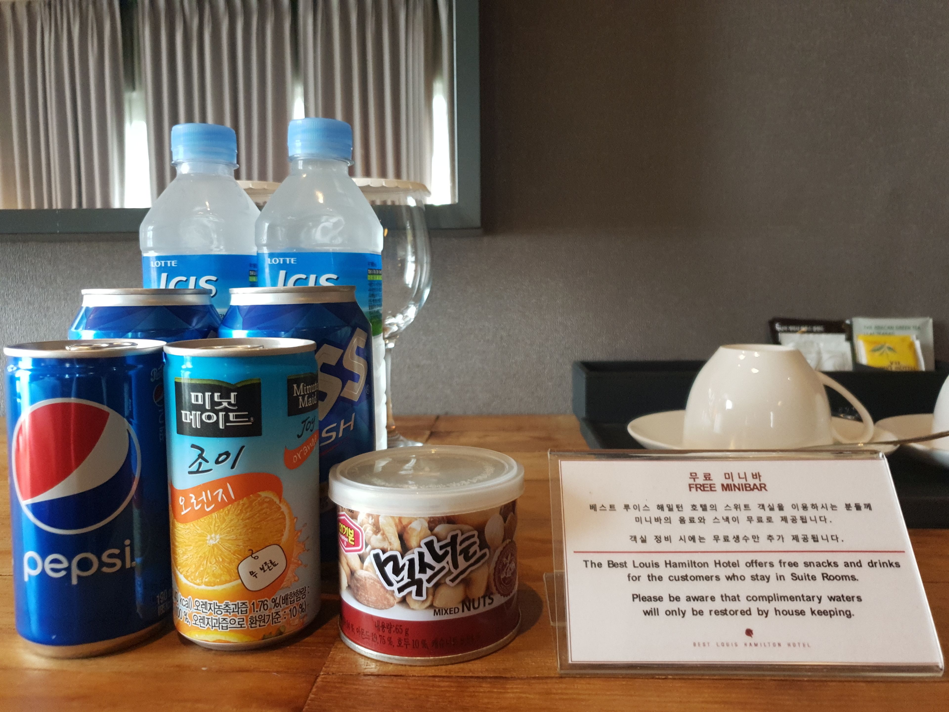 A table with various beverages including Pepsi, Lotte Ices, and mixed nuts. There is also a sign about complimentary snacks and drinks at the Best Louis Hamilton Hotel.