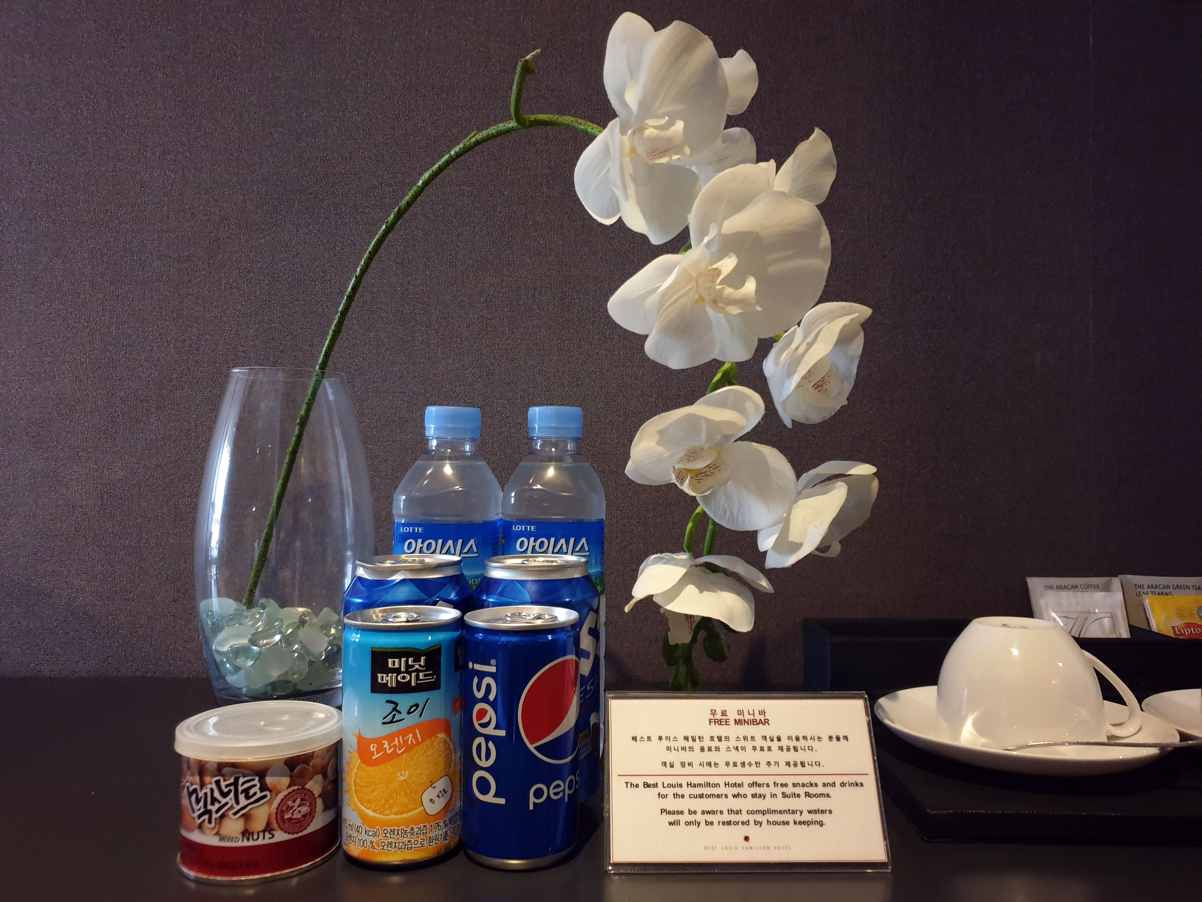 A minibar setup with bottled water, soda, and a can of nuts, along with a vase of white flowers and a plate with a cup.