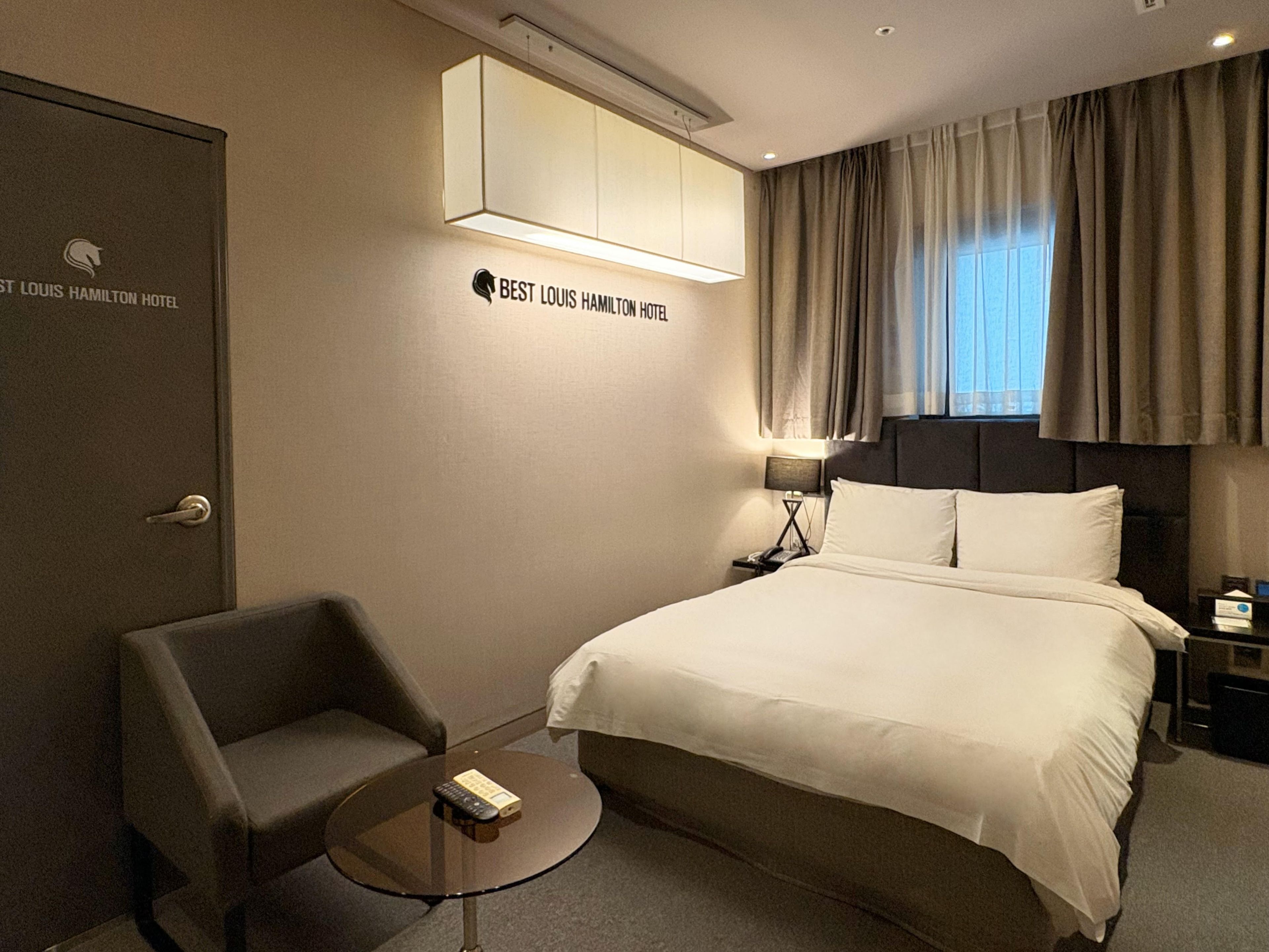 A hotel room with a bed, a chair, a small table, a nightstand, and a wall-mounted air conditioning unit.