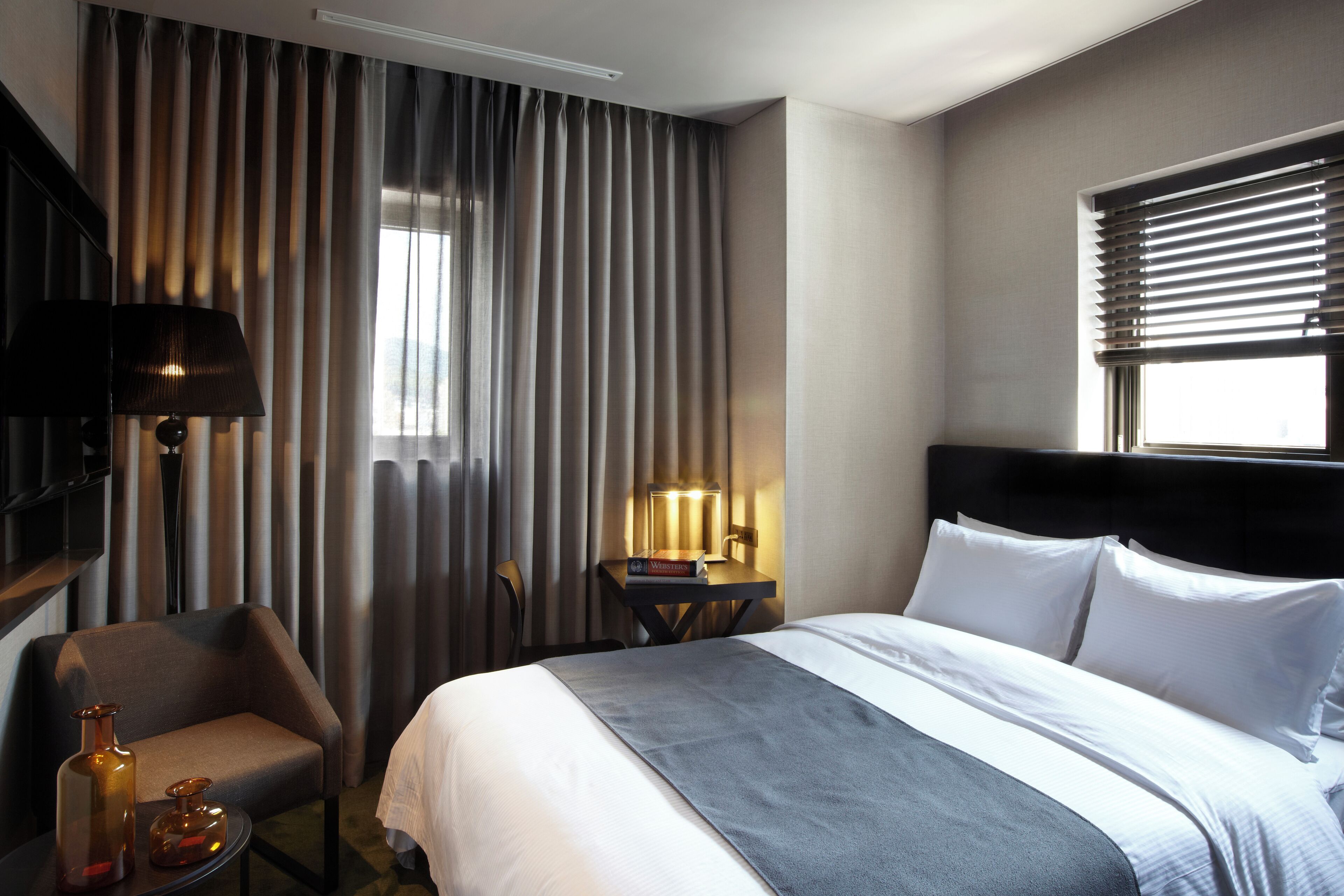 A hotel room with a large bed, a desk with a lamp, a chair, and a window with blinds.