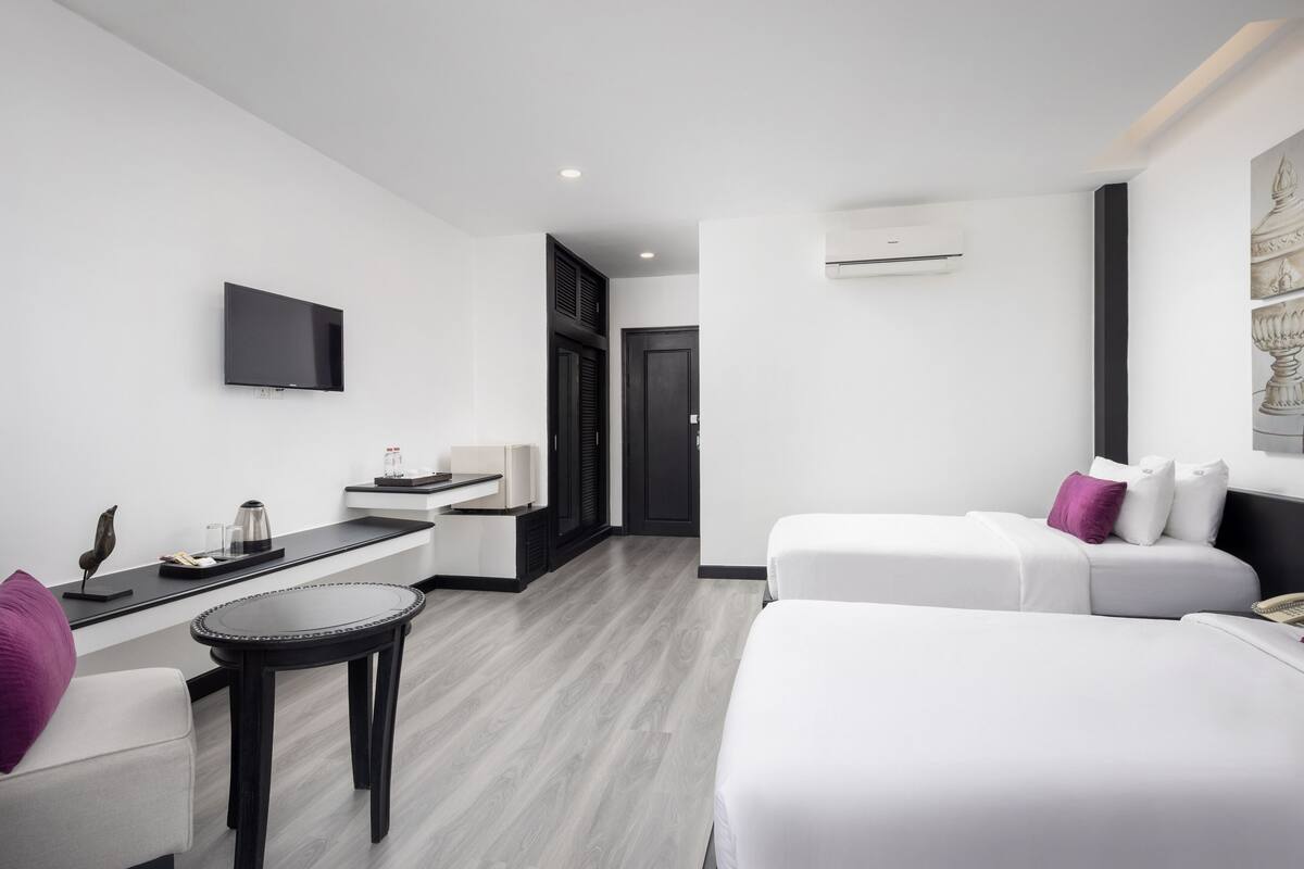 Ein modernes Hotelzimmer mit Flachbildfernseher, einem kleinen runden Tisch, zwei Betten mit weißer Bettwäsche und violetten Kissen, einer Kaffeemaschine und einem schwarzen Schreibtisch mit Flachbildfernseher.