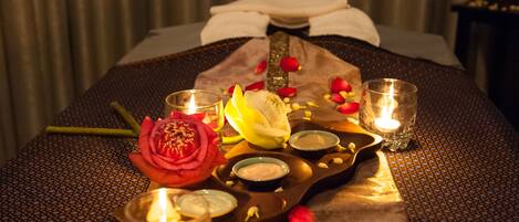 A neatly arranged bed with rose petals, candles, and a tray of snacks.