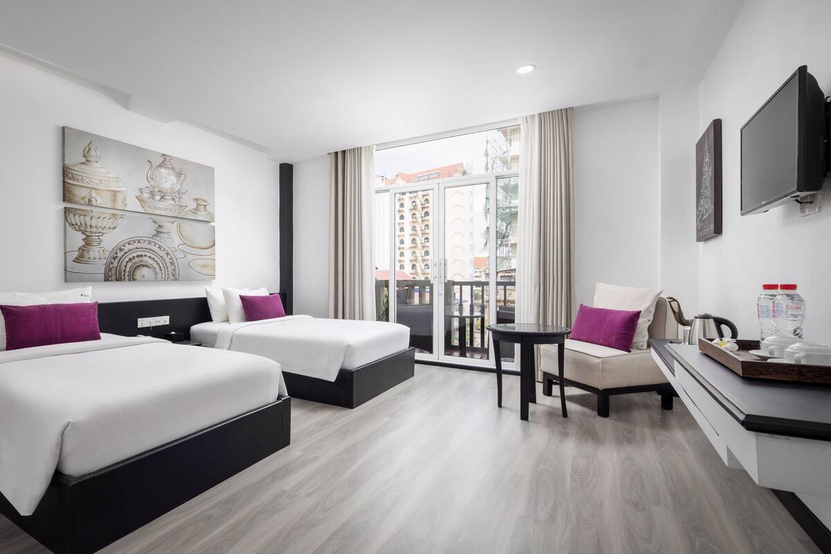 Ein Hotelzimmer mit zwei Betten, einem kleinen Tisch, einem Sessel, einem Fernseher und einem Balkon mit Blick auf Gebäude.