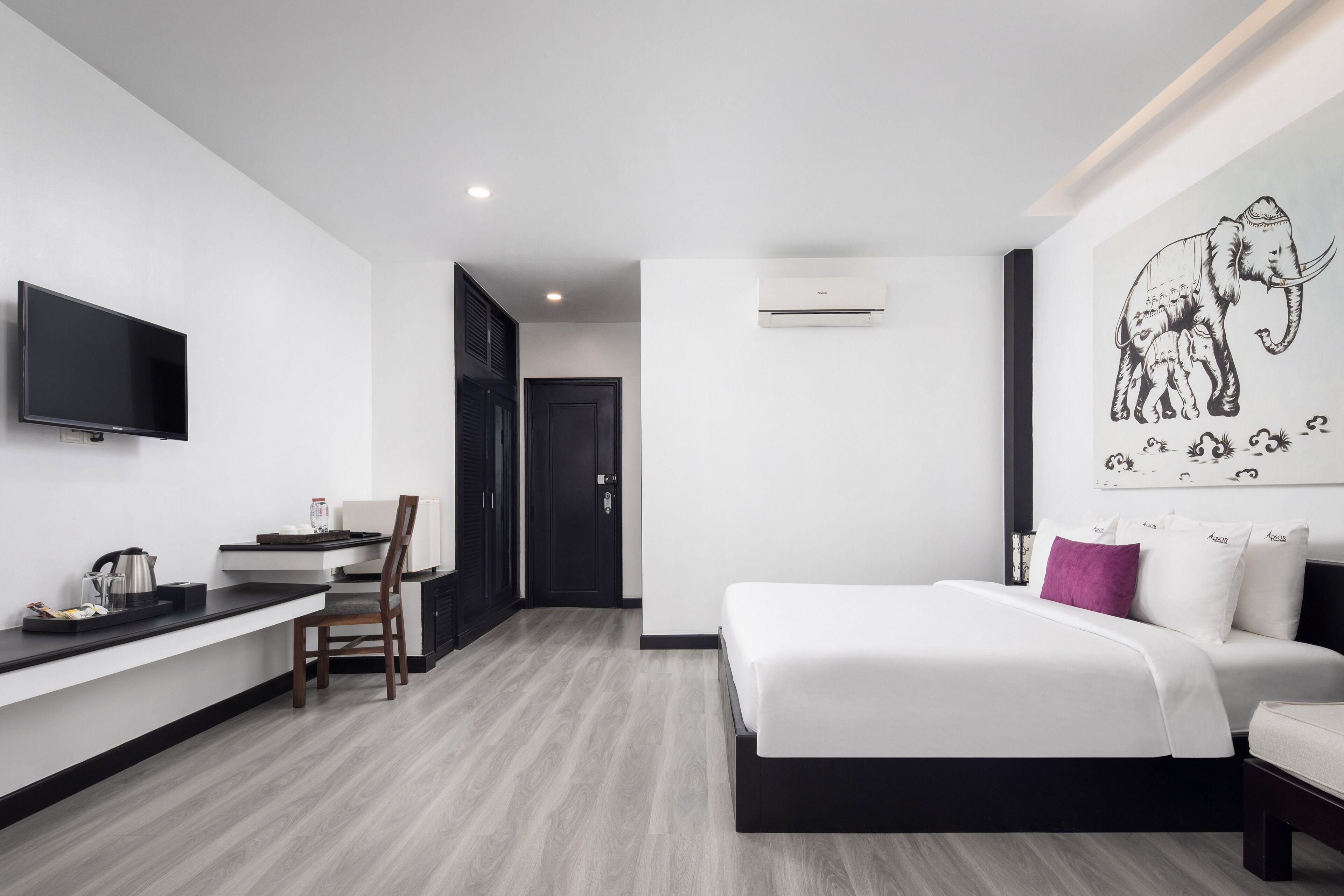 Camera d'albergo moderna con TV a schermo piatto, una scrivania con sedia, un letto con biancheria bianca e un cuscino viola, e un murale raffigurante elefanti.
