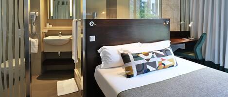 Habitación de hotel moderna con una cama grande, una mesita de noche, un escritorio, un baño con lavamanos y espejo, y vistas al exterior.