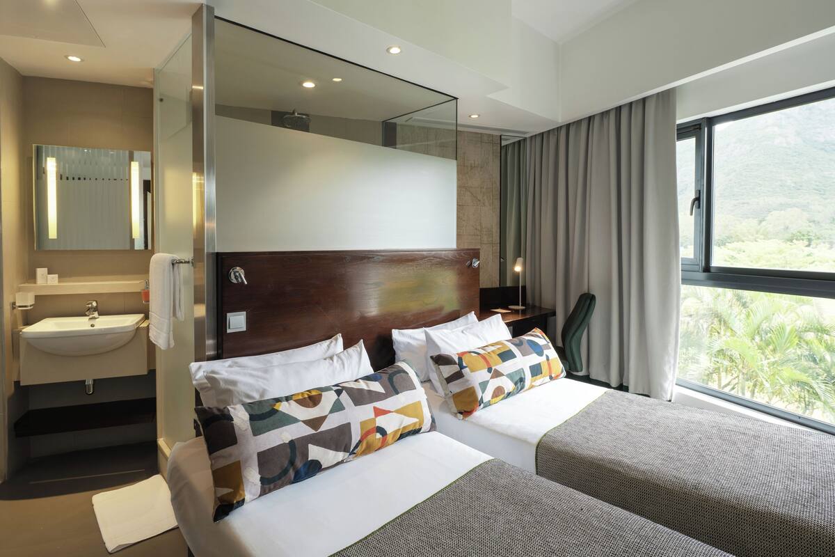 Une chambre d’hôtel moderne avec un grand lit, une table de chevet, un miroir et une salle de bain équipée d’un lavabo, offrant une vue sur la verdure par la fenêtre.
