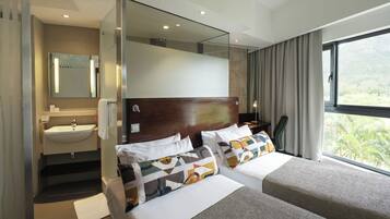 Una habitación de hotel moderna con una cama grande, una mesita de noche, un espejo, un baño con lavamanos y vistas a zonas verdes a través de la ventana.