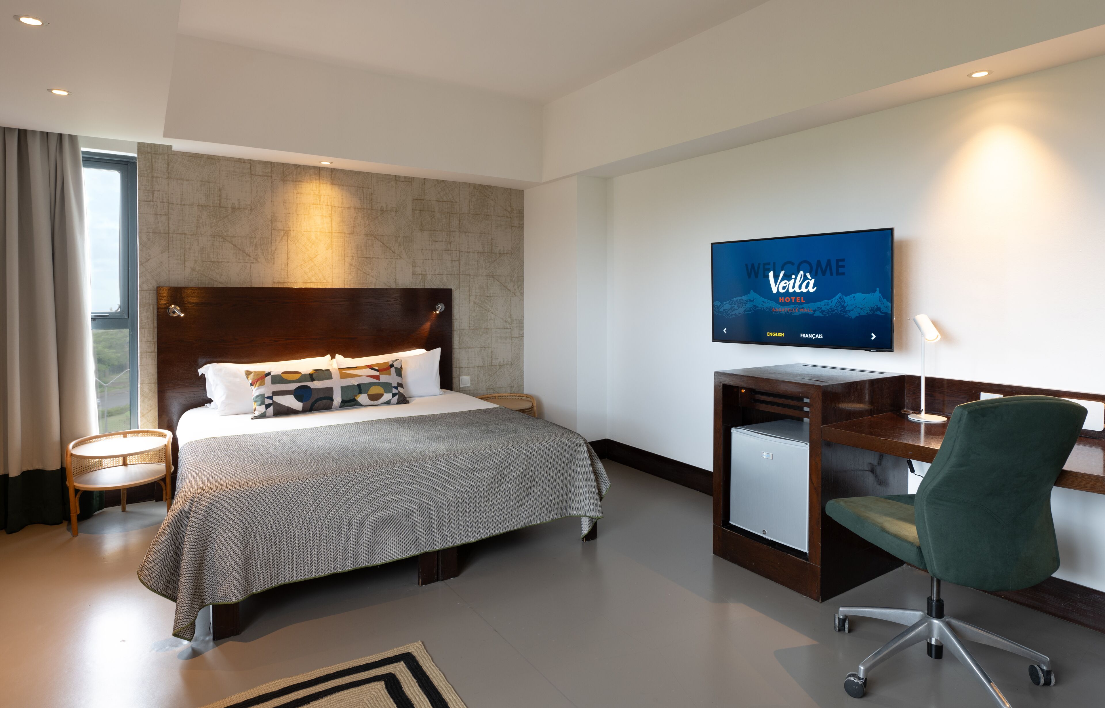 A modern hotel room with a large bed, a desk, and a television displaying the hotel's name.