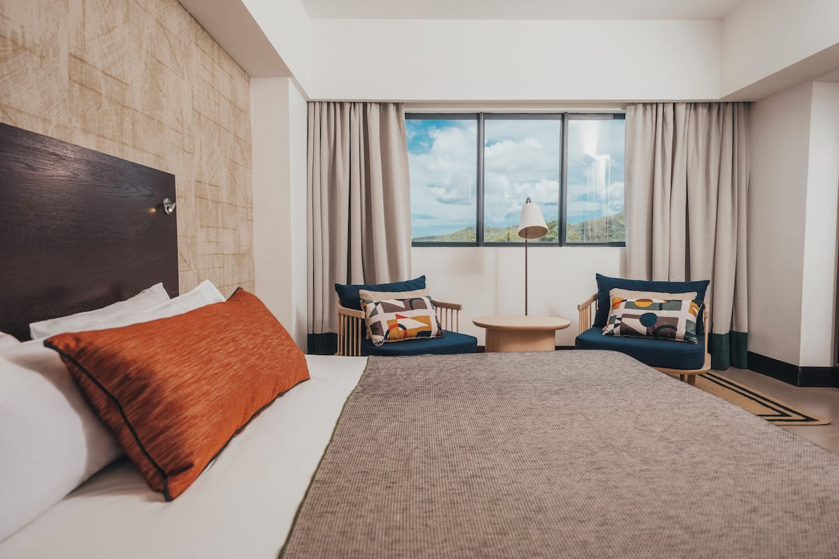 Une chambre d’hôtel avec un grand lit, deux fauteuils, une petite table et une vue sur l’extérieur par une fenêtre.