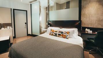 Ein modernes Hotelzimmer mit einem großen Bett, einem Schreibtisch, einem Sessel, einem Spiegel und einem durch eine Glastür einsehbaren Badezimmer.