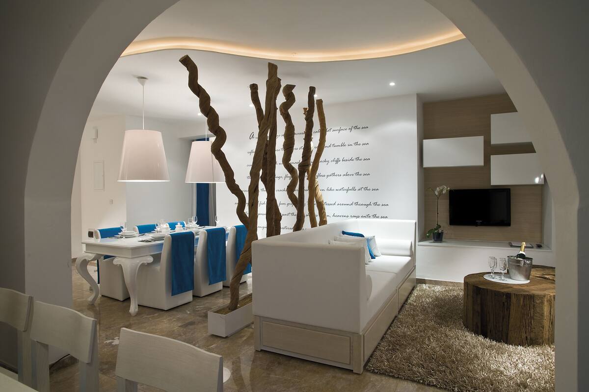 Een moderne woonkamer met een witte bank, een eettafel met blauwe stoelen, een houten salontafel en een aan de muur bevestigde televisie.