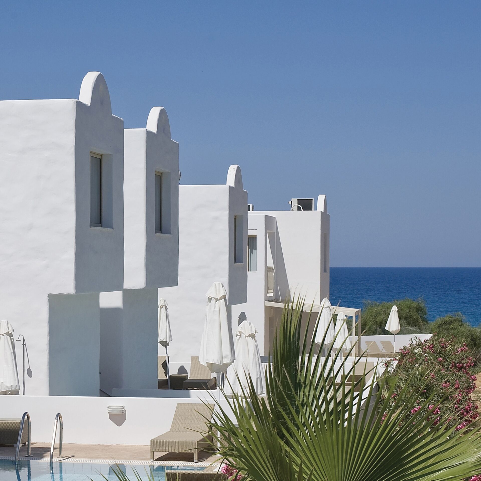 A white building with a pool and umbrellas, overlooking the sea.