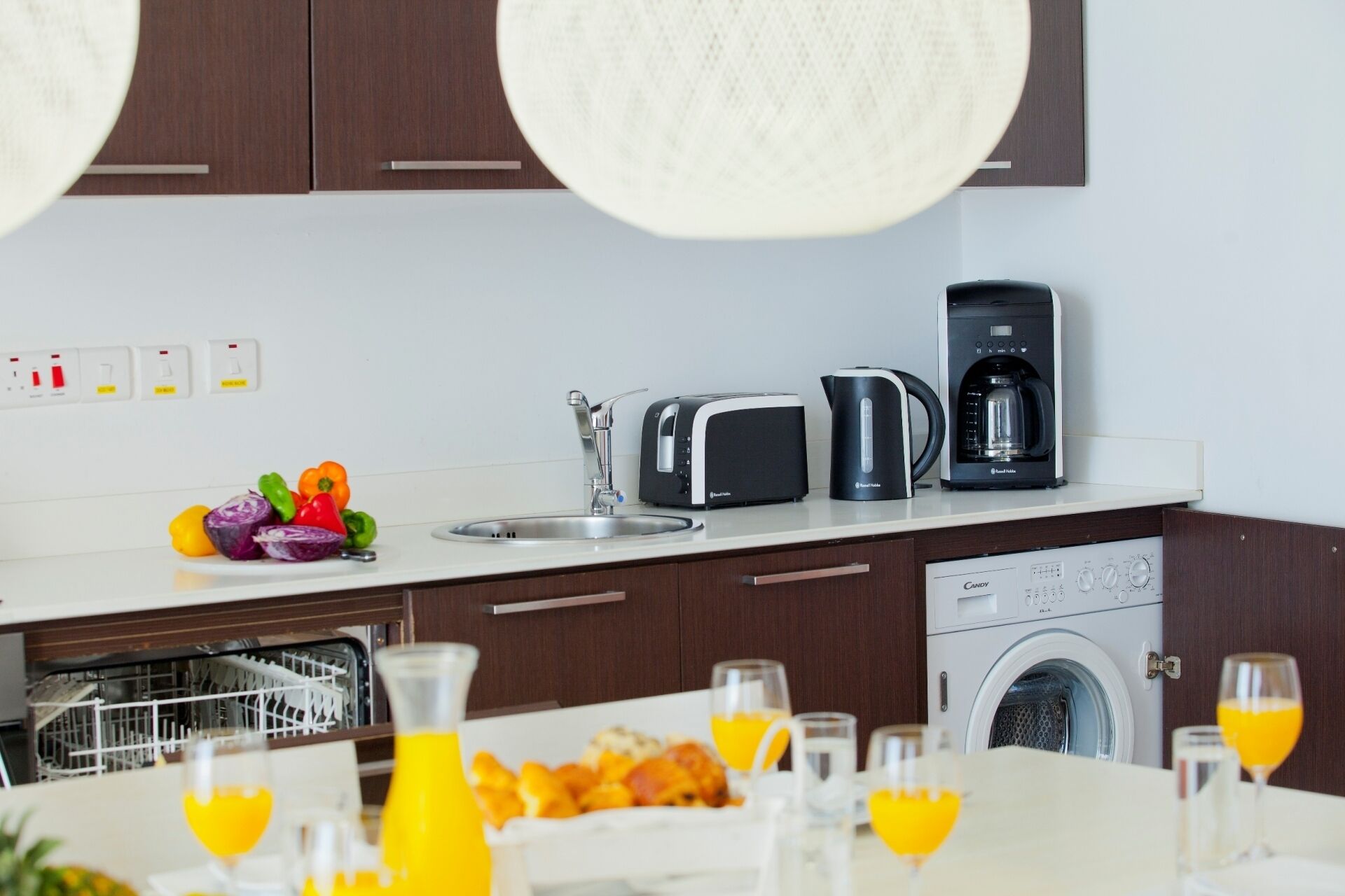 A modern kitchen with a sink, toaster, kettle, and coffee maker. There are fruits and vegetables on the counter, and a dishwasher and washing machine in the background.