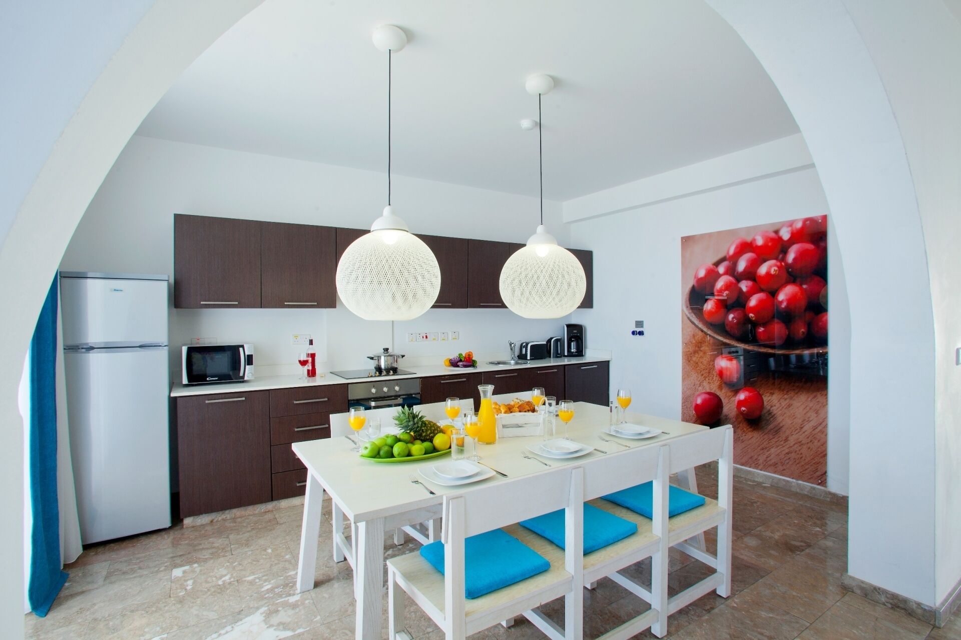 Een moderne keuken met witte kastjes, een eettafel met blauwe kussens, en een grote fruitschaal aan de muur.