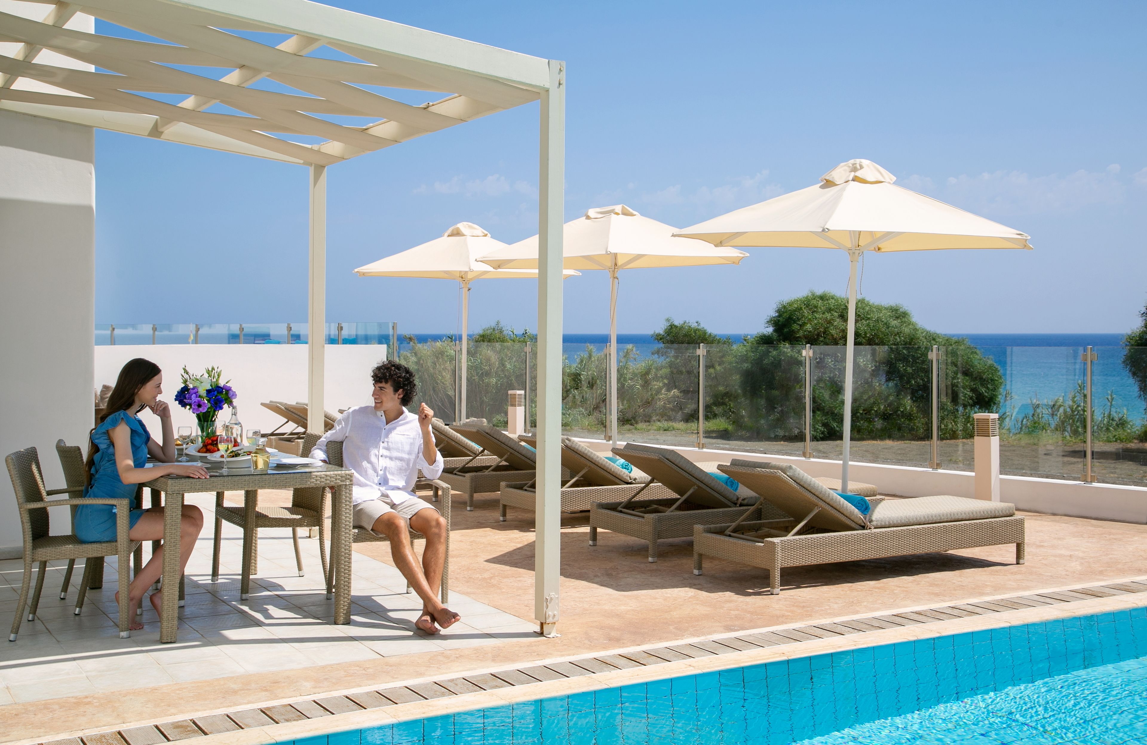 A poolside area with a table and chairs, lounge chairs, and umbrellas.