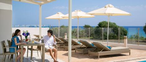A poolside area with a table and chairs, lounge chairs, and umbrellas.