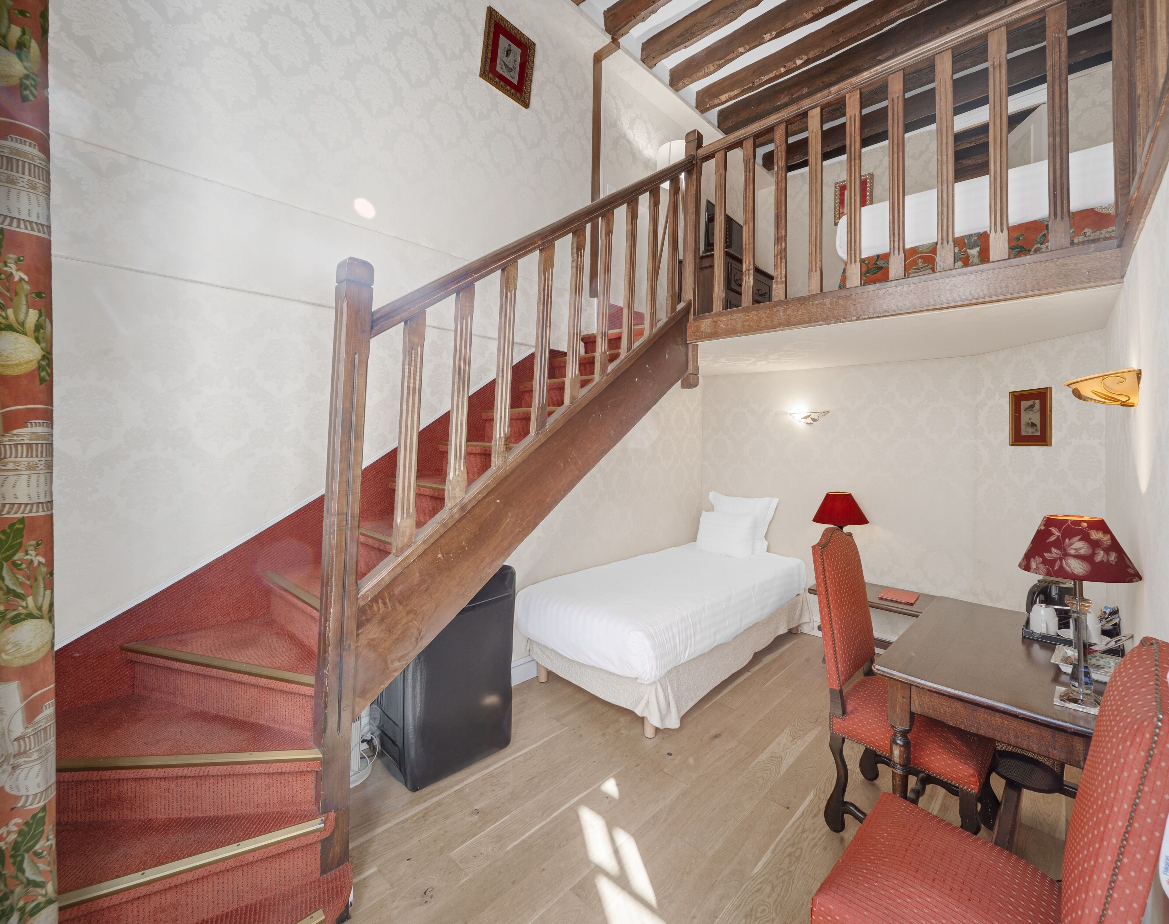 A bedroom with a wooden staircase, a bed, a desk with a chair, and a small table with a lamp.