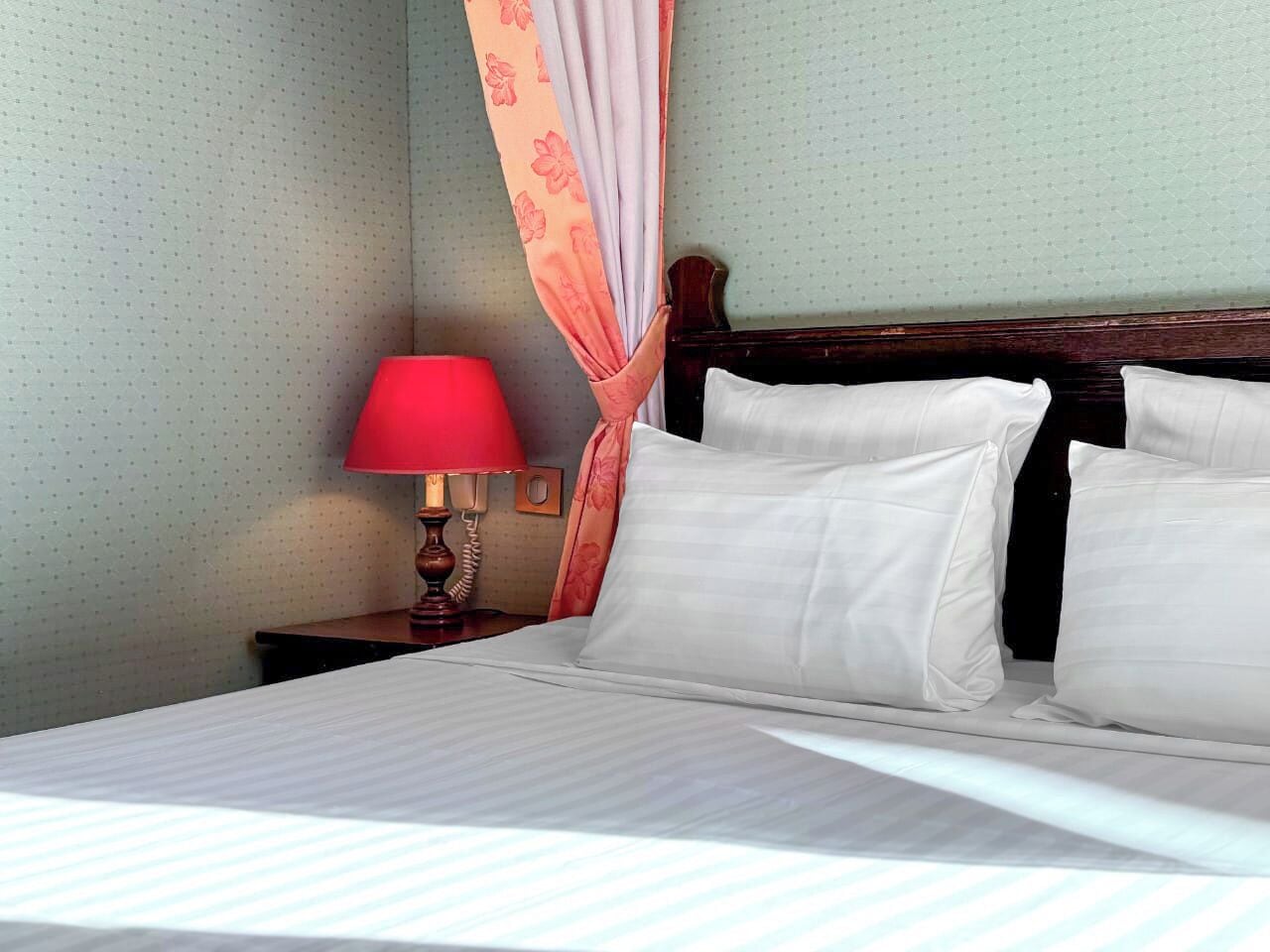 A neatly made bed with white linens and pillows, a wooden headboard, a red lamp, and a floral curtain on a wall with a polka dot pattern.