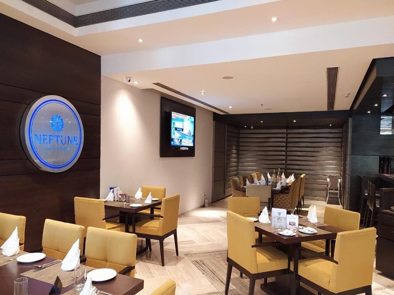 A modern hotel lobby with a Neptune sign, dining area with yellow chairs, and a TV screen.