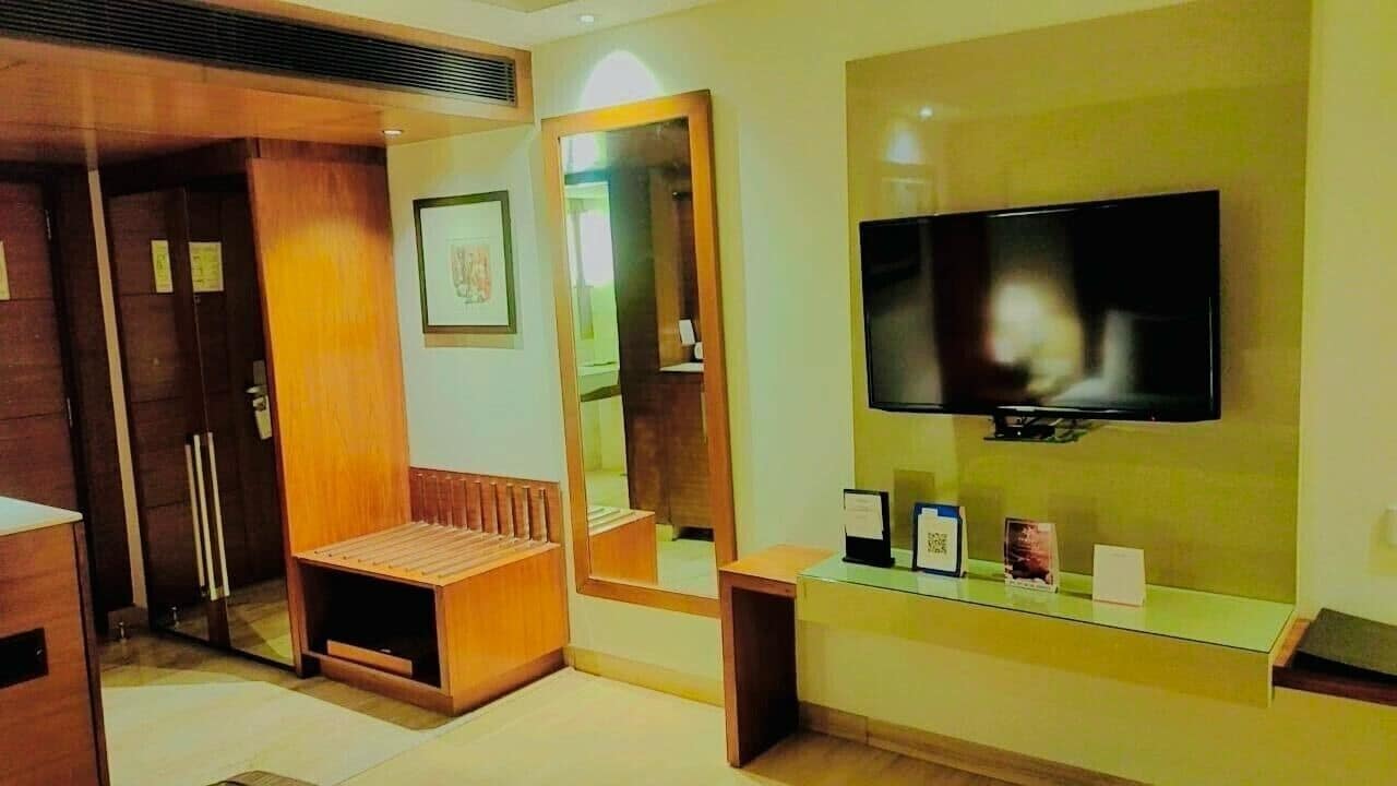A hotel room with a wooden bench, a large mirror, a television mounted on the wall, and a small table with a few items on it.