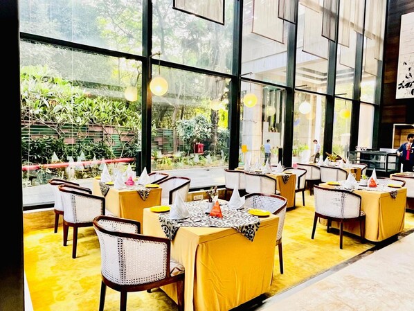 A restaurant with yellow tablecloths, wicker chairs, and large windows offering a view of greenery.