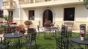 Patio esterno con diversi tavoli e sedie rotondi, circondato da piante in vaso e un prato curato.