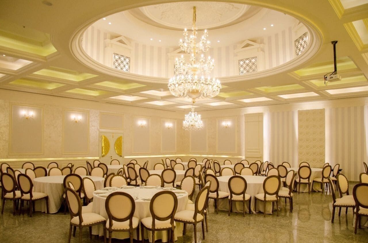 A banquet hall with round tables and chairs, a large chandelier, and a high ceiling with decorative moldings.