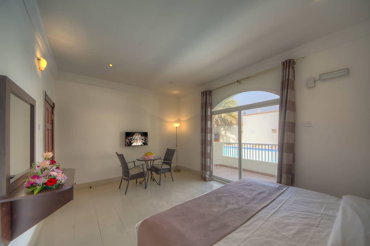 A hotel room with a bed, a desk with a chair, a small table with a lamp, and a large window with curtains.